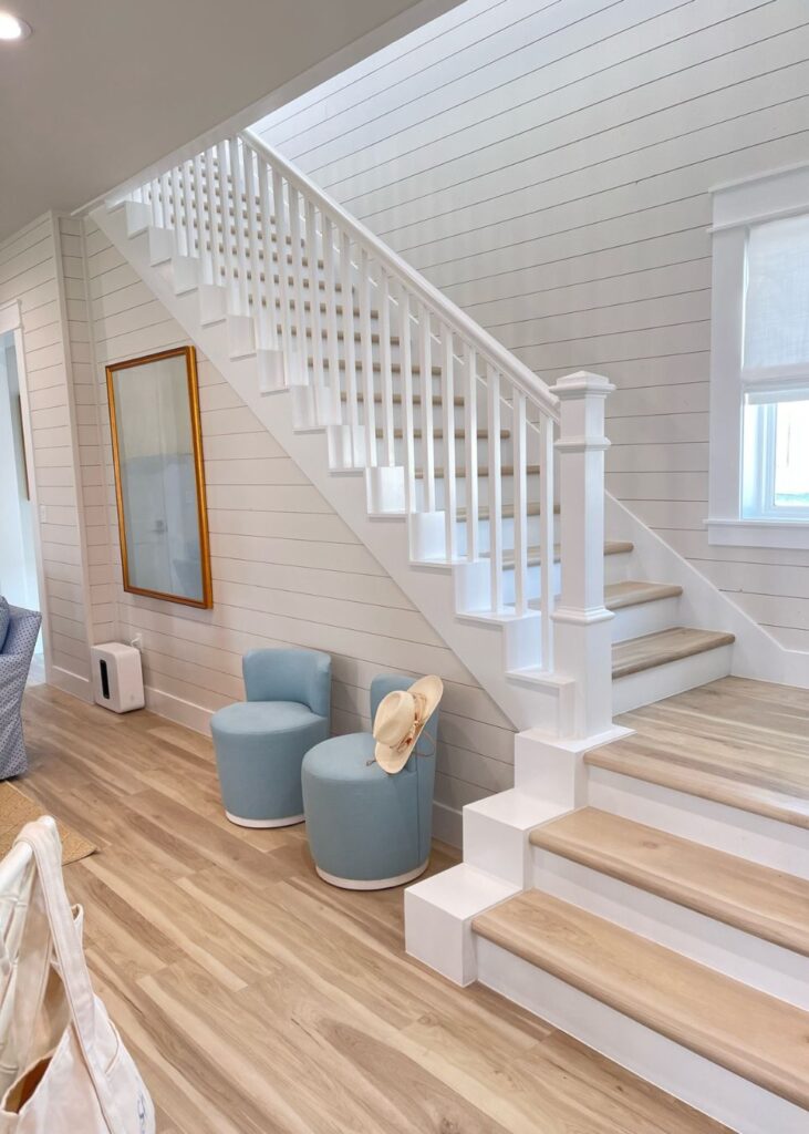A bright, airy room designed by an interior designer Boerne TX features light wood floors, white shiplap walls, a wooden staircase with white rails, two light blue stools, a tan hat, a framed mirror, and a small air purifier.