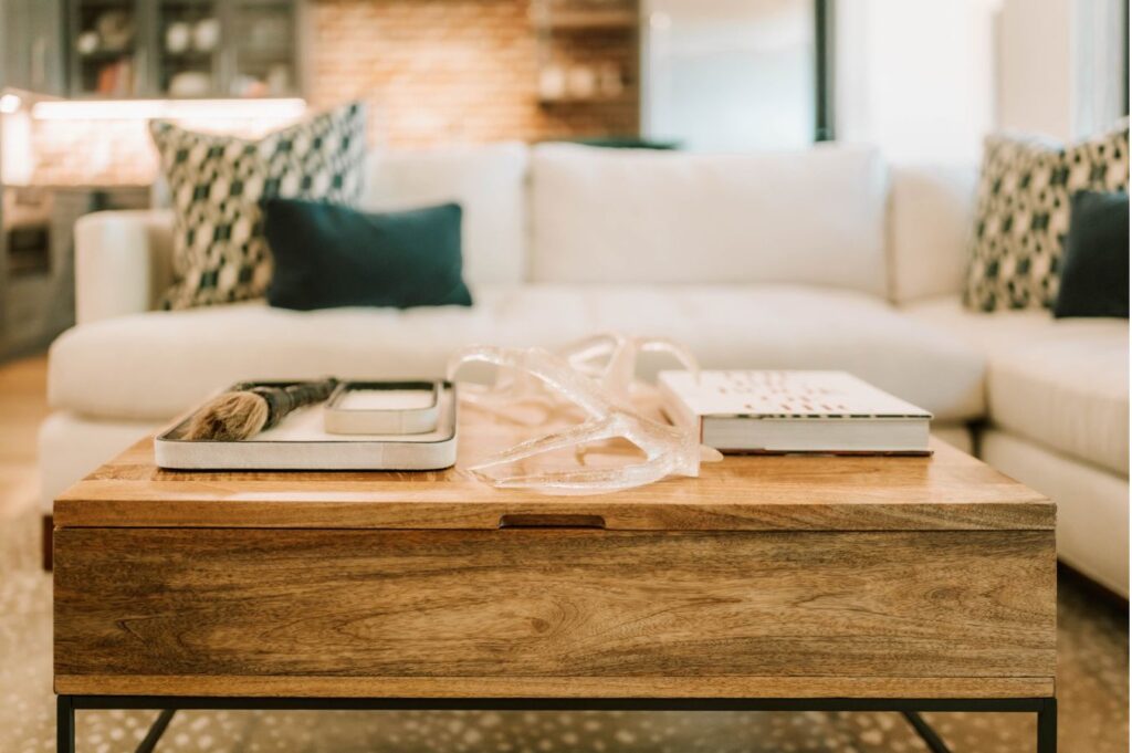 A wooden coffee table with decorative items, styled by an interior designer Boerne TX, sits in front of a white sofa with patterned and solid pillows in a bright, modern living room.