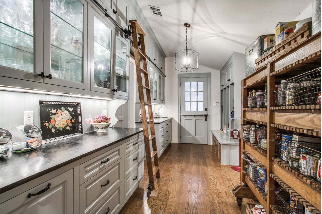 A spacious pantry with white cabinets, glass-fronted uppers, a wooden countertop, and a rolling ladder—expertly arranged by an interior designer Boerne TX—features wooden shelves of canned goods and hardwood flooring leading to a windowed door.