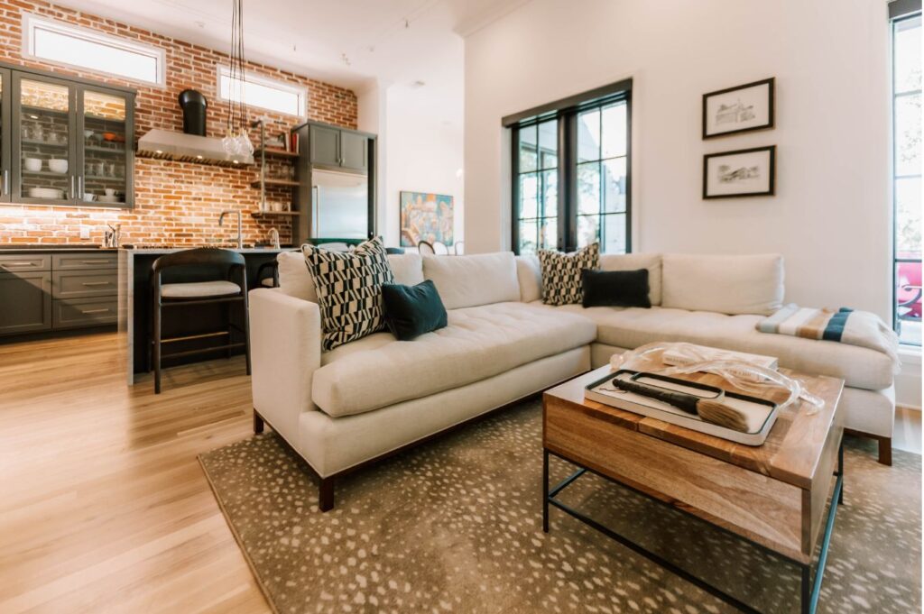 A modern living room designed by an interior designer Boerne TX features a white sectional sofa, patterned cushions, a wooden coffee table, and an open kitchen with brick walls. Large windows flood the space with natural light.