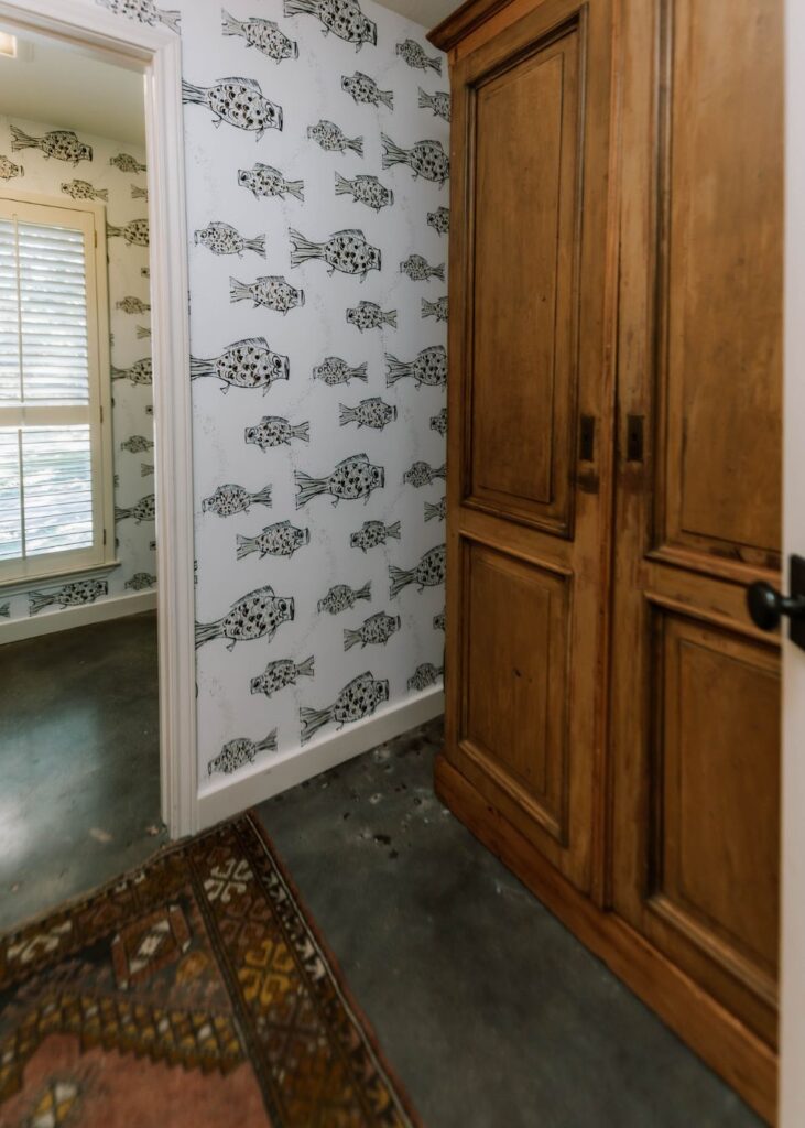 A hallway with fish-patterned wallpaper, a large wooden wardrobe, and a colorful rug on concrete flooring, designed by an expert in interior design Boerne TX. A doorway opens to a room with shuttered windows.