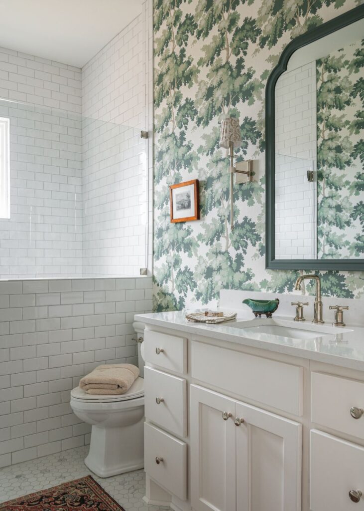 Bright bathroom with white subway tile walls, leafy green wallpaper, a white vanity with silver fixtures, a large mirror, and neatly folded towels—perfect inspiration for fresh bathroom design ideas.