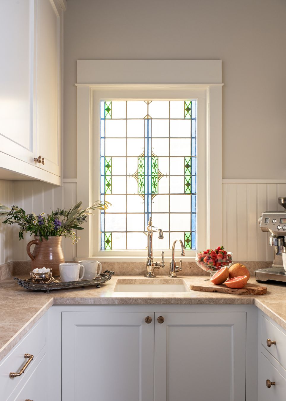 Bright kitchen with white cabinets, a marble countertop, and a sink beneath a decorative stained glass window. Styled by an interior designer Boerne TX, the counter features coffee, flowers, fruit, and a coffee machine.