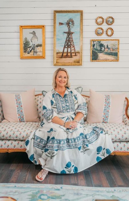 A woman in a long, patterned dress sits on a vintage-style sofa with decorative pillows. Designed by an interior decorator Boerne, the cozy room features shiplap walls and framed paintings of a windmill and bird for a warm, rustic vibe.