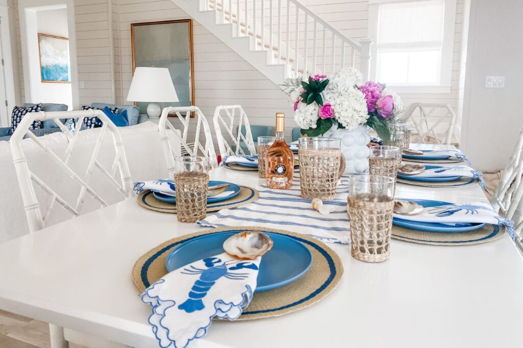 A bright dining table set for six with blue lobster-themed napkins, blue plates, and a floral centerpiece creates a beachy vibe, styled by an expert interior decorator Boerne in this inviting coastal-style room.