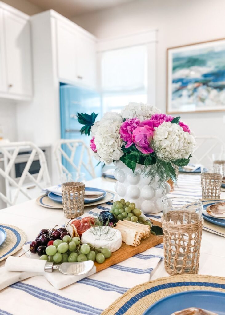 A dining table set with blue and white plates, wicker glasses, a cheese and fruit board, and a white vase of pink and white flowers sits in the center. The background reveals a bright kitchen styled by an interior design company in Boerne, TX.
