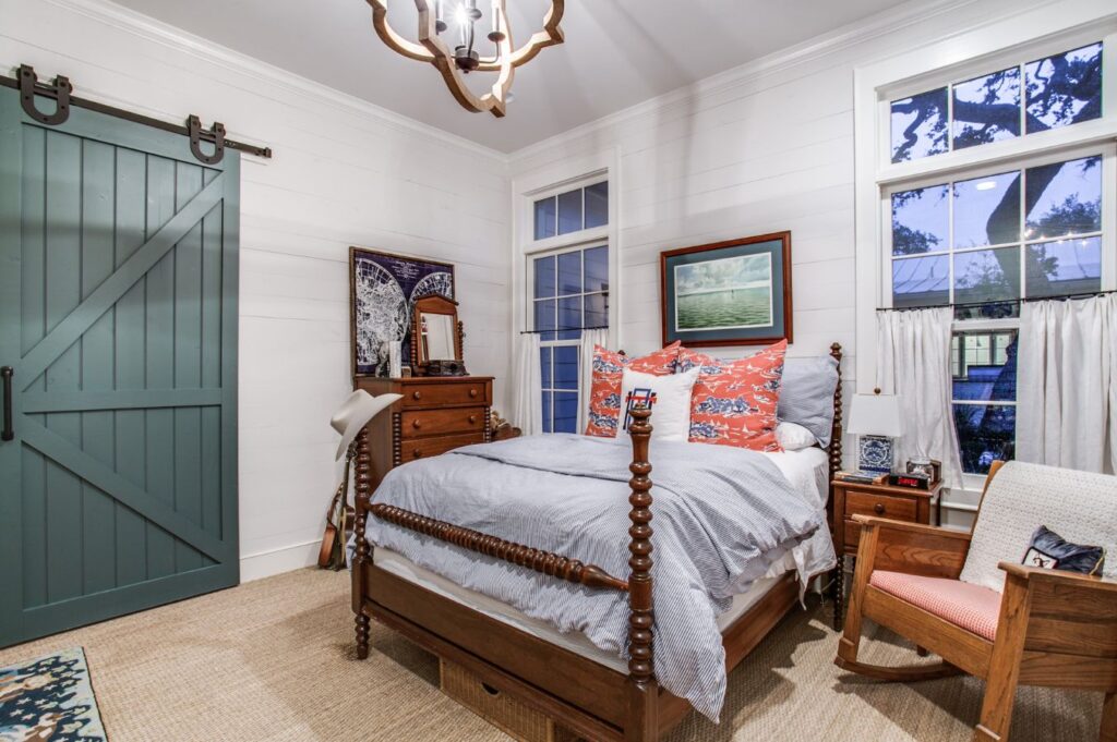 A cozy bedroom with a wooden bed, blue bedding, and patterned pillows. Designed by an interior decorator Boerne, the space features a rocking chair, wooden furniture, teal sliding barn door, and large windows that let in natural light.