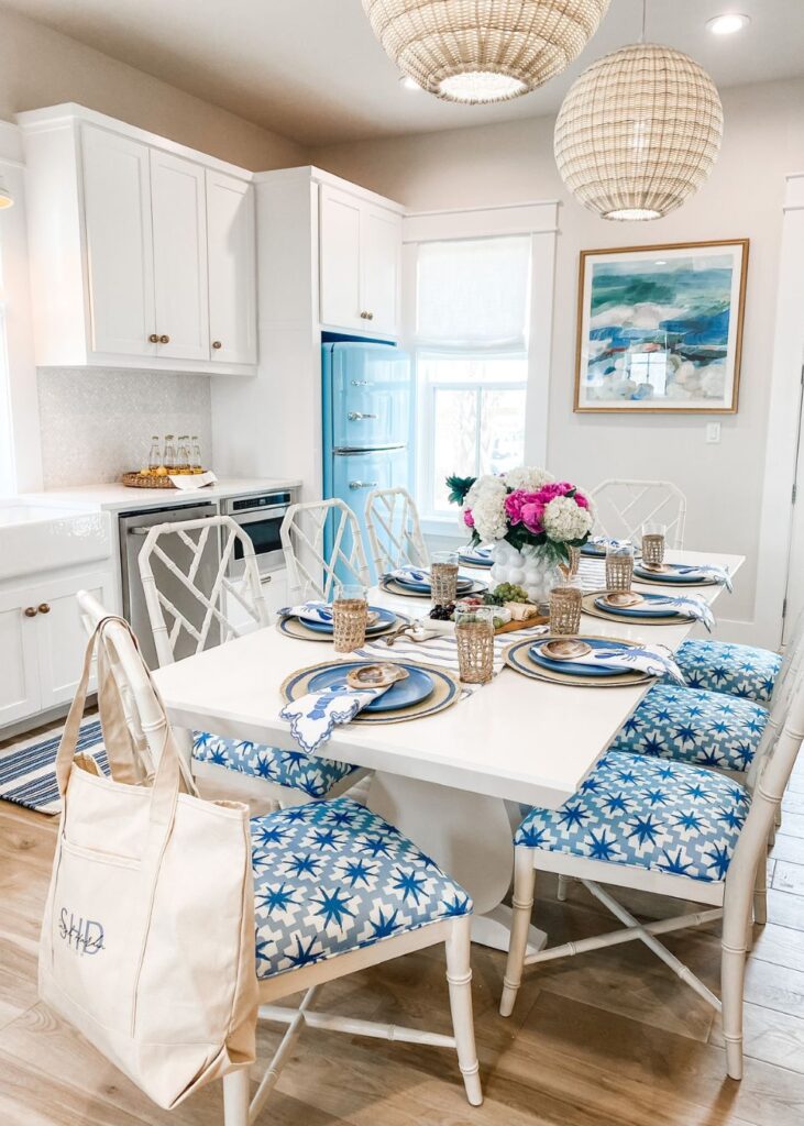 A bright dining area with a white table set for six, blue and white star-patterned chairs, gold-accented plates, floral centerpiece, and woven pendant lights—styled by an expert interior designer Boerne TX—in a modern white kitchen.