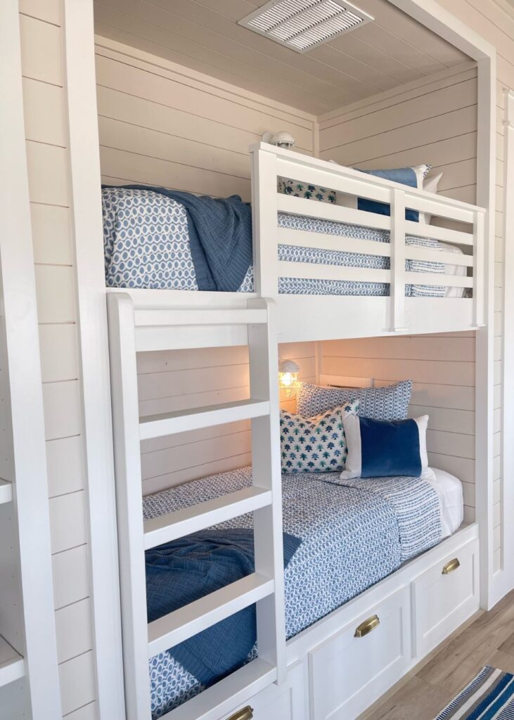 White built-in bunk beds with blue and white bedding, shiplap walls, and brass drawer pulls below. Designed by an interior designer in Boerne, TX, each bunk features a blue blanket, patterned pillows, and a small light above the lower bed.