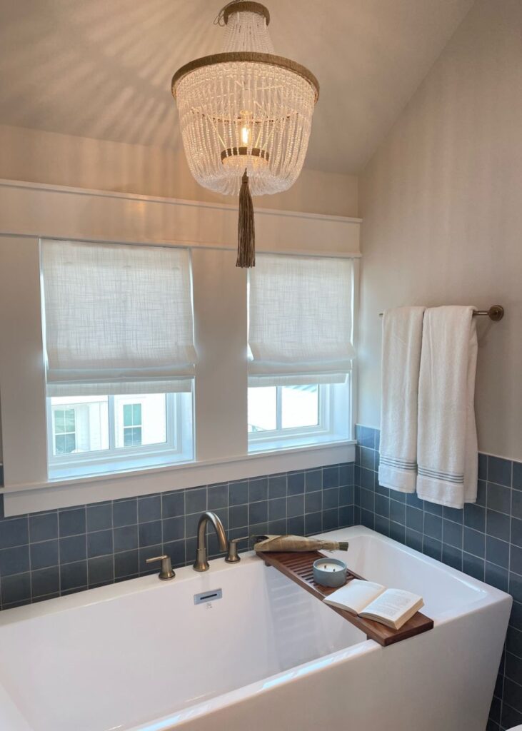A modern bathroom, styled by an interior designer Boerne TX, features a white bathtub with a wooden bath tray holding a book and cup. A beaded chandelier hangs above, while blue tiled walls and roman shades brighten the space. White towels hang nearby.