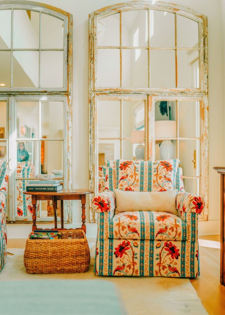 A cozy armchair with a colorful floral and striped pattern sits beside a wooden side table topped with books, styled by an interior designer Boerne TX, all set in front of large, rustic window-pane mirrors.