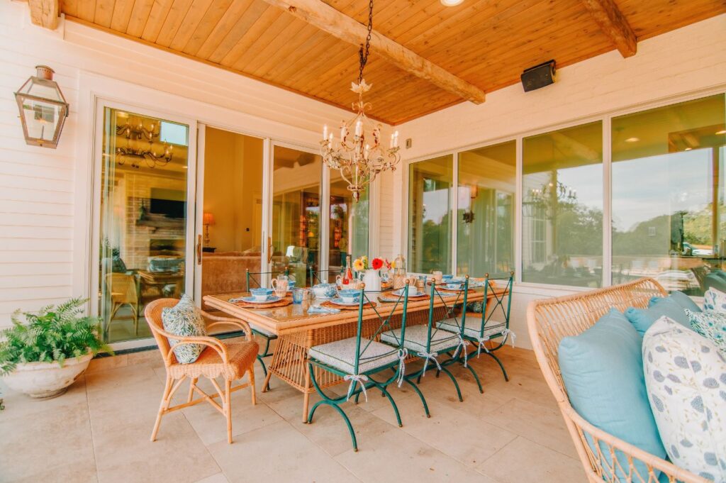 A cozy outdoor patio designed by an interior designer Boerne TX, featuring a long dining table with chairs and a cushioned bench. A chandelier hangs from the wooden ceiling, and large windows offer beautiful views of the greenery outside.