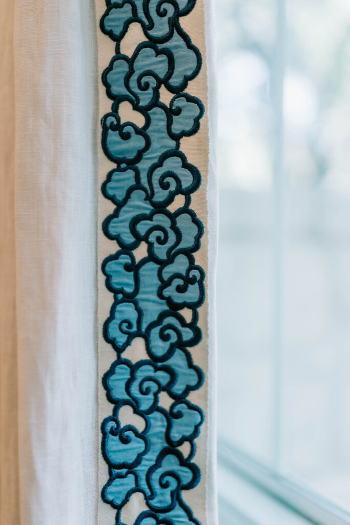 A white curtain features a vertical trim with blue, cloud-like embroidered patterns. Perfect for a serene touch, it’s a choice often recommended by an interior designer Boerne TX for rooms filled with soft natural light.