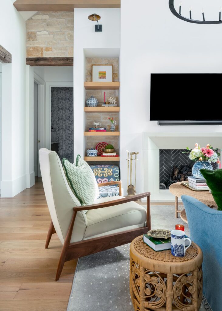 A modern living room with wooden floors, a white armchair with green cushions, and built-in shelves exudes style—showcasing the expertise of an interior designer Boerne TX. A wall-mounted TV crowns the fireplace, while a wicker side table holds a coffee mug and book.