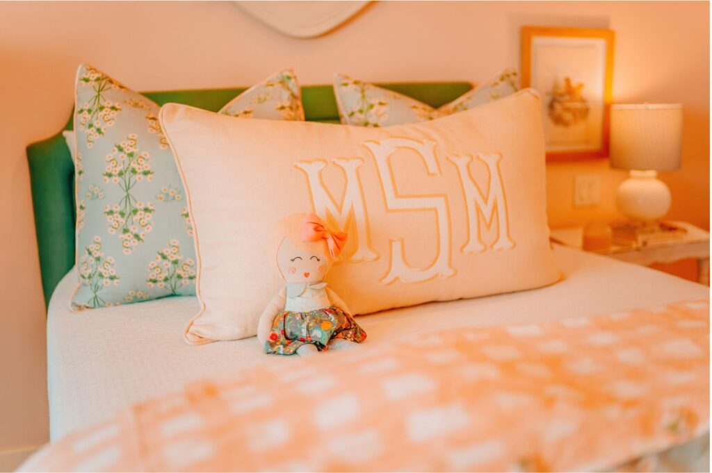 A neatly made bed with a large monogrammed pillow reading MSM, floral accent pillows, and a doll with a pink bow sits in front. Styled by an expert interior designer Boerne TX, the softly lit room features a lamp and framed picture on the bedside table.