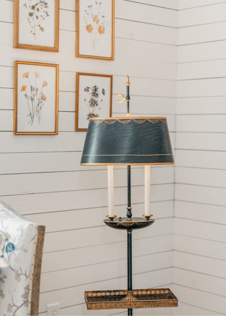 A black and gold vintage lamp with two candle-like bulbs sits in front of a white shiplap wall adorned with framed dried flowers, showcasing the elegant touch of an interior designer Boerne TX. A floral-patterned chair appears on the left.
