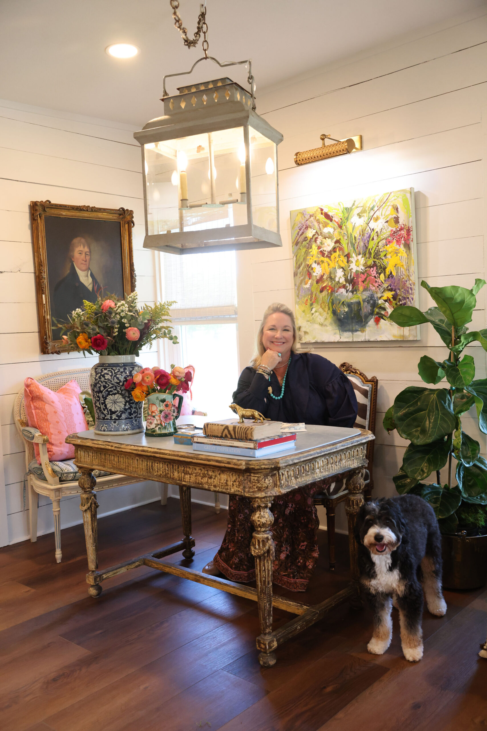 Sarah Hardin seated at an antique gilt writing table in her studio, her dog beside her, beneath a metal lantern
