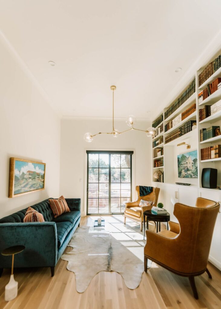 Bright, stylish living room by an interior design company in Boerne, TX, featuring a blue sofa, tan leather armchairs, cowhide rug, modern chandelier, artwork, and large glass doors opening to a sunny patio.