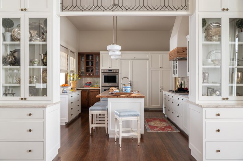White kitchen with glass-front upper cabinets, a painted island, open shelving and a vent hood, designed by Sarah Hardin Design.