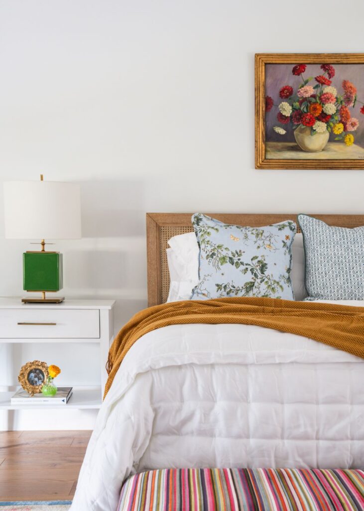 A cozy bedroom styled by an interior design company in Boerne, TX, features floral and blue cushions, a white comforter, mustard blanket, and a colorful bench. A green lamp and flower painting add charm to this inviting retreat.