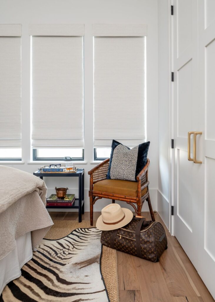 A cozy bedroom corner curated by an interior designer Boerne TX features a rattan chair with pillows, a side table with books and decor, a zebra-print rug, plus a Louis Vuitton duffel bag and straw hat on the wooden floor near the bed.