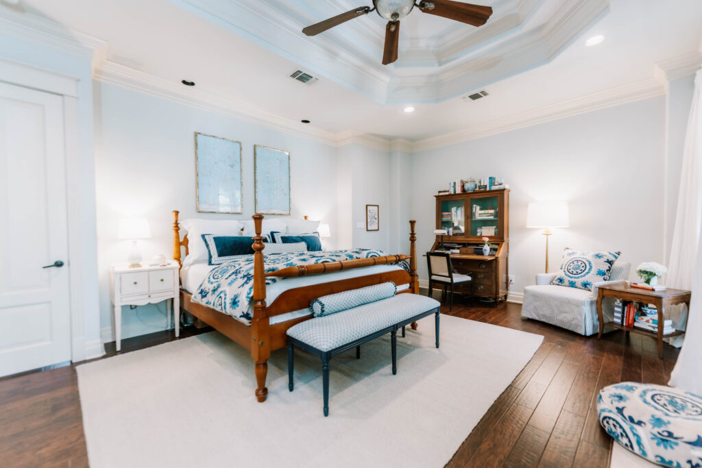 Bright bedroom with a wooden bed, blue and white bedding, nightstands, a bench, writing desk with lamp, armchair, hardwood floors, and ceiling fan—expertly styled by an interior designer Boerne TX for comfort and elegance.