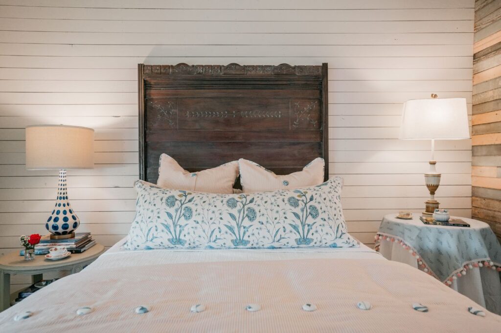 A cozy bedroom features a wooden headboard, floral pillows, a white bedspread, and two bedside tables with lamps. Designed by an interior designer in Boerne, TX, the white shiplap walls create a serene retreat with books, flowers, and a teacup.