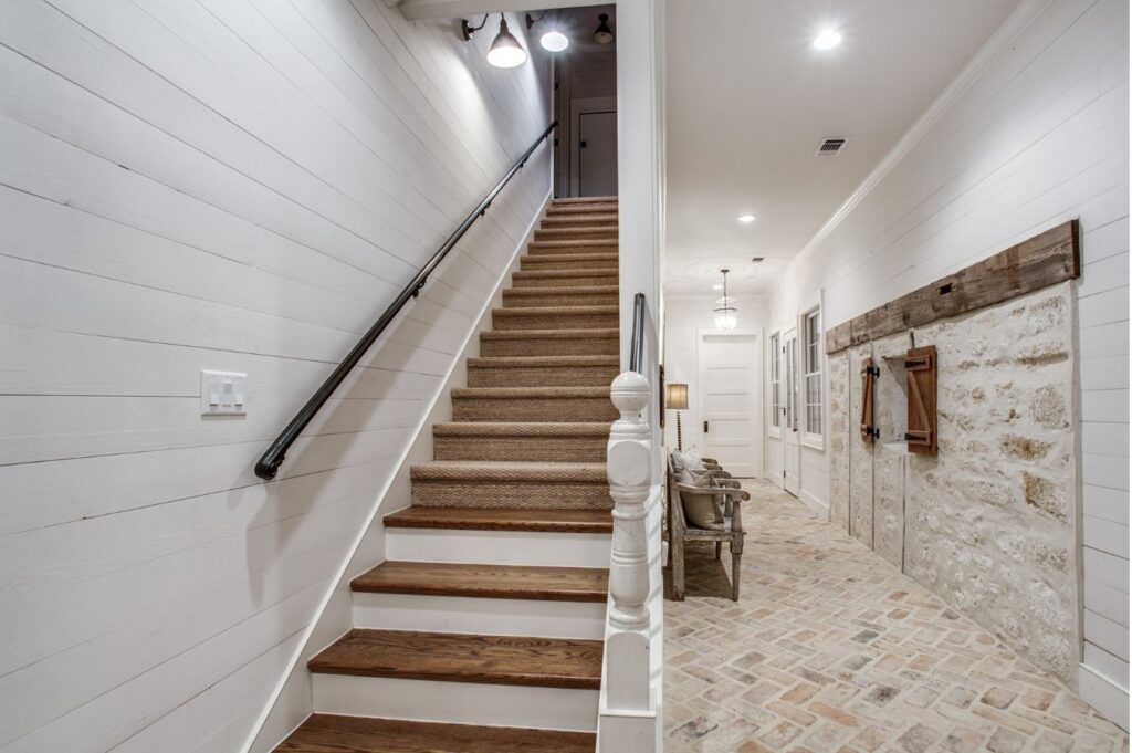 A farmhouse-style hallway, designed by an interior decorator Boerne, features brick flooring, shiplap walls, a rustic sliding barn door, and a staircase with a dark handrail and carpeted steps. The space is brightly lit with ceiling lights.