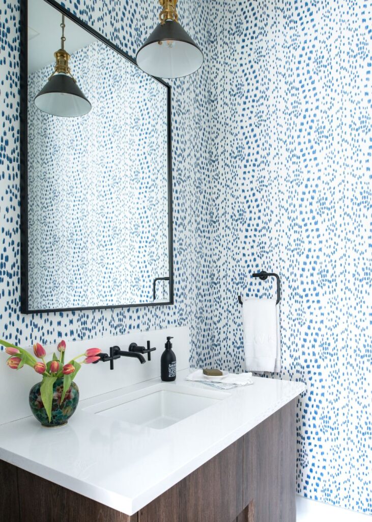 Modern bathroom by an interior design company in Boerne, TX, featuring blue and white dotted wallpaper, a rectangular mirror with black frame, white countertop with dark wood vanity, black wall-mounted faucet, floral vase, towel, and pendant lights.