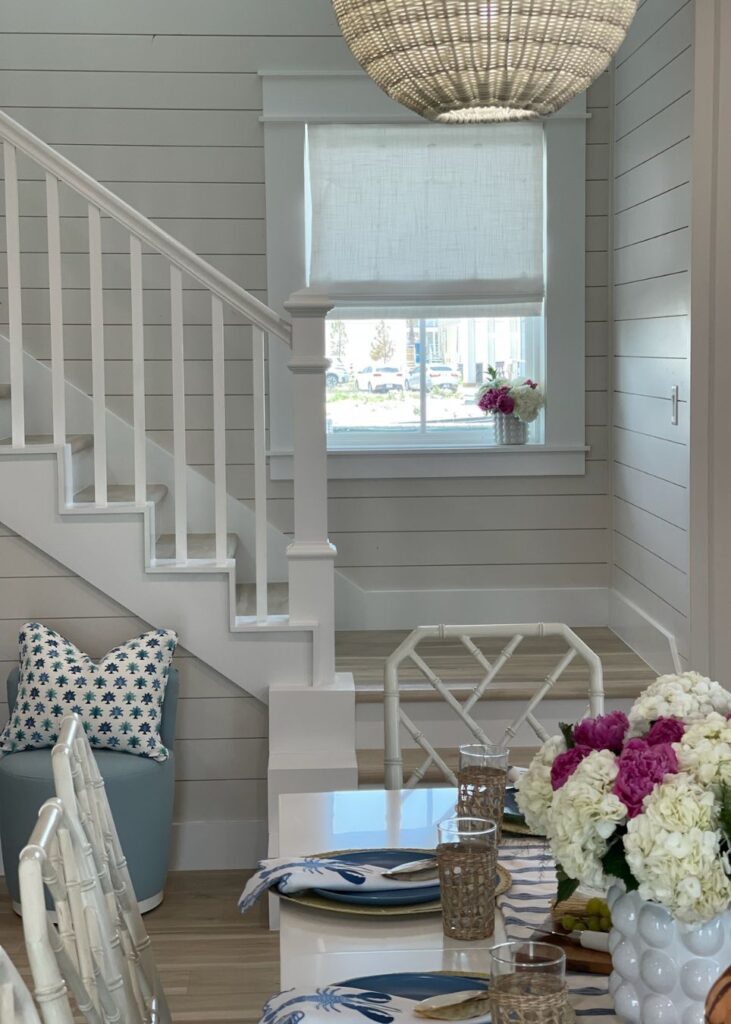 A bright, coastal-style dining area by a top interior designer in Boerne, TX, featuring white shiplap walls, a staircase with white railings, a set table for four, and blue starfish pillows for a charming beachy touch.