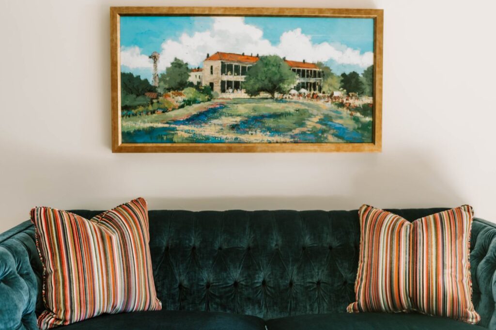 A framed landscape painting hangs on a white wall above a dark green tufted sofa with two striped pillows. Styled by an interior designer Boerne TX, the space blends color and comfort for a welcoming atmosphere.