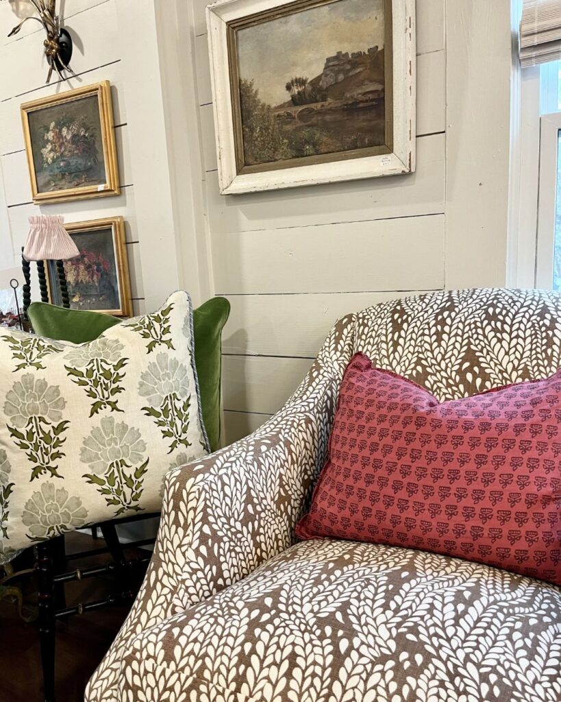 A cozy corner curated by an interior designer Boerne TX features a patterned armchair, pink decorative pillow, and white floral pillow. Three framed paintings on beige paneled walls add a charming vintage touch to the space.