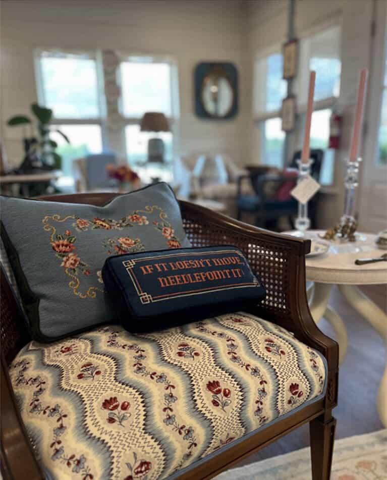 A decorative chair with floral upholstery holds two cushions—one embroidered, one reading IF IT DOESN’T MOVE, NEEDLEPOINT IT—in a cozy, sunlit living room styled by an interior designer Boerne TX with pastel decor and candles.