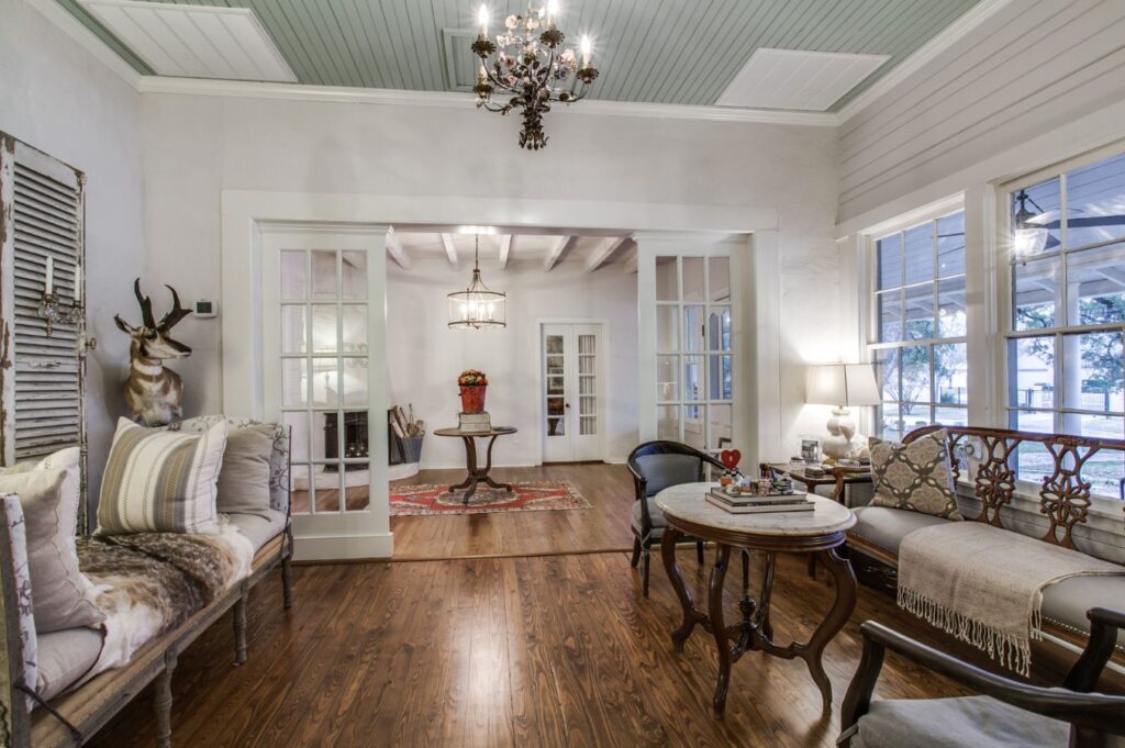A cozy, vintage-style living room by an interior designer in Boerne, TX, featuring wooden floors, antique furniture, a chandelier, and a wall-mounted deer head. French doors open to a bright room with classic furnishings and a patterned rug.