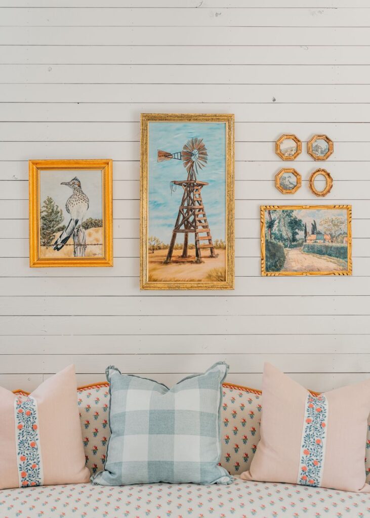 A cozy sofa with patterned pillows sits against a white shiplap wall adorned by five framed pictures, styled by an expert interior decorator Boerne, adding a touch of personalized charm to the space.
