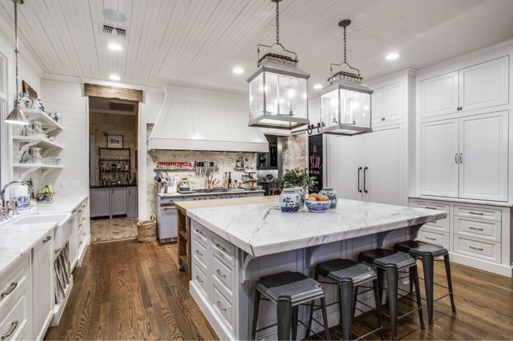 A spacious, elegant kitchen designed by a top interior decorator Boerne features white cabinetry, marble countertops, a large island with stools, pendant lights, farmhouse sink, wood floors, and open shelves. A pantry is visible through a doorway.