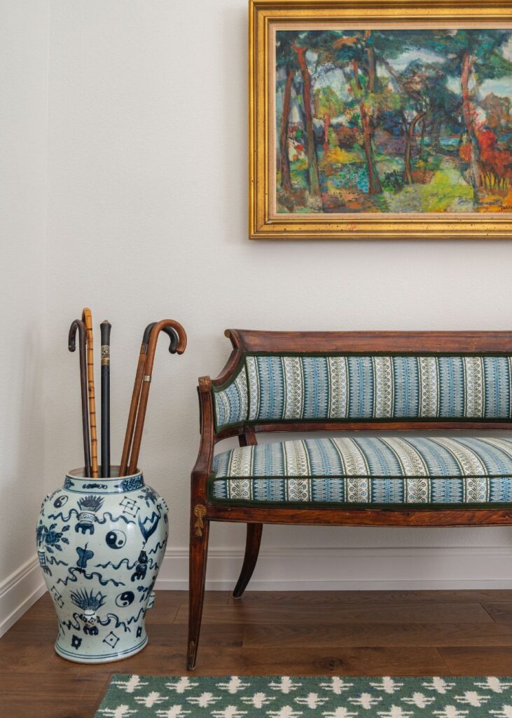 A vintage wooden bench with blue patterned upholstery sits beside a large ceramic vase holding three canes. Above, a colorful framed painting of a forest scene hangs on a white wall. A green patterned rug is on the floor.