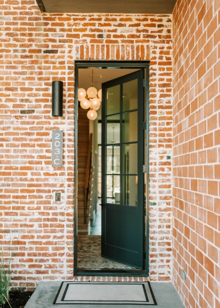 The image shows a partially open black-framed glass door on a red brick house. A modern black wall light and a vertical address plaque with 203C are next to the door, reflecting the style an interior design company Boerne TX might recommend.