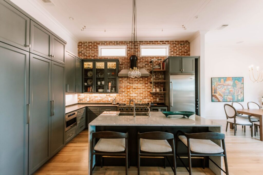 Modern kitchen with black cabinetry, brick backsplash, stainless steel appliances, and a large island with three chairs. Pendant lights hang above the island. Designed by an interior designer Boerne TX, dining area visible in the background.