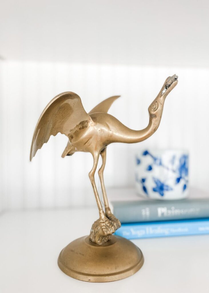 A brass crane figurine with outstretched wings stands on a round base, styled by an interior designer Boerne TX; behind it, a blue and white cup and two stacked books with blue covers rest on a white surface.