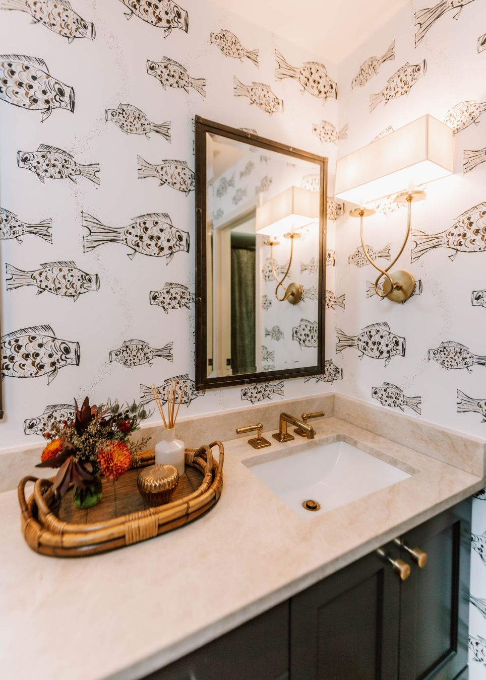 Bathroom with a marble countertop, gold fixtures, a rectangular mirror, and fish wallpaper. Styled by an interior designer Boerne TX, it features wall-mounted lights and a wicker tray with flowers and a candle on the counter.