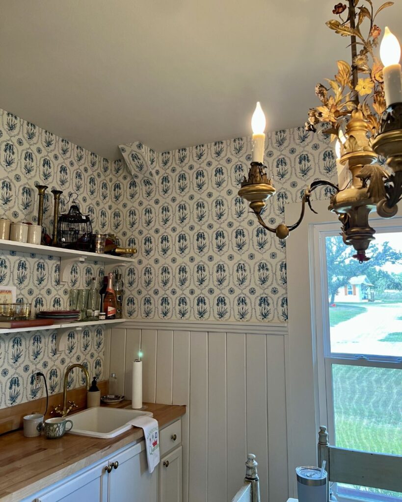 A cozy kitchen with blue and white patterned wallpaper, wooden countertops, open shelves, and an ornate chandelier. Sunlight streams in—a charming retreat styled by a leading interior design company Boerne TX.
