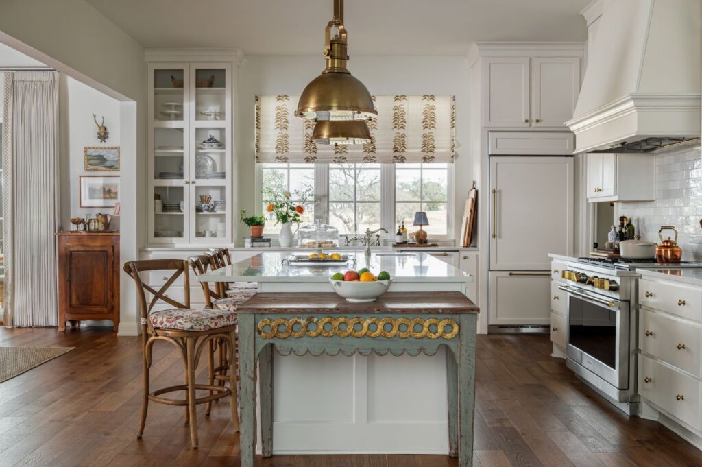 A bright kitchen designed by a top kitchen designer in Boerne features white cabinets, a central island with ornate trim, wooden chairs, brass pendant lights, marble countertops, and large windows with patterned curtains that welcome natural light.