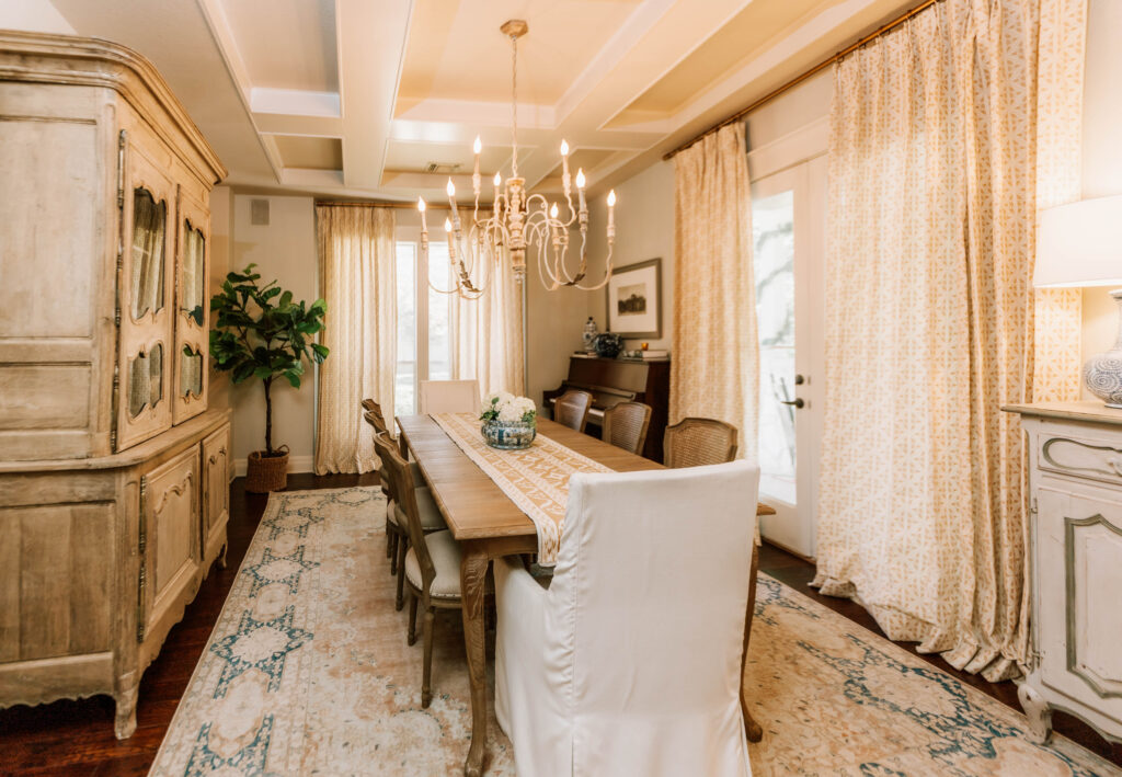 Elegant dining room with a long wooden table, eight chairs, chandelier, potted plant, antique cabinets, patterned rug, and light curtains over tall windows—perfect inspiration from an interior design company Boerne TX residents trust.