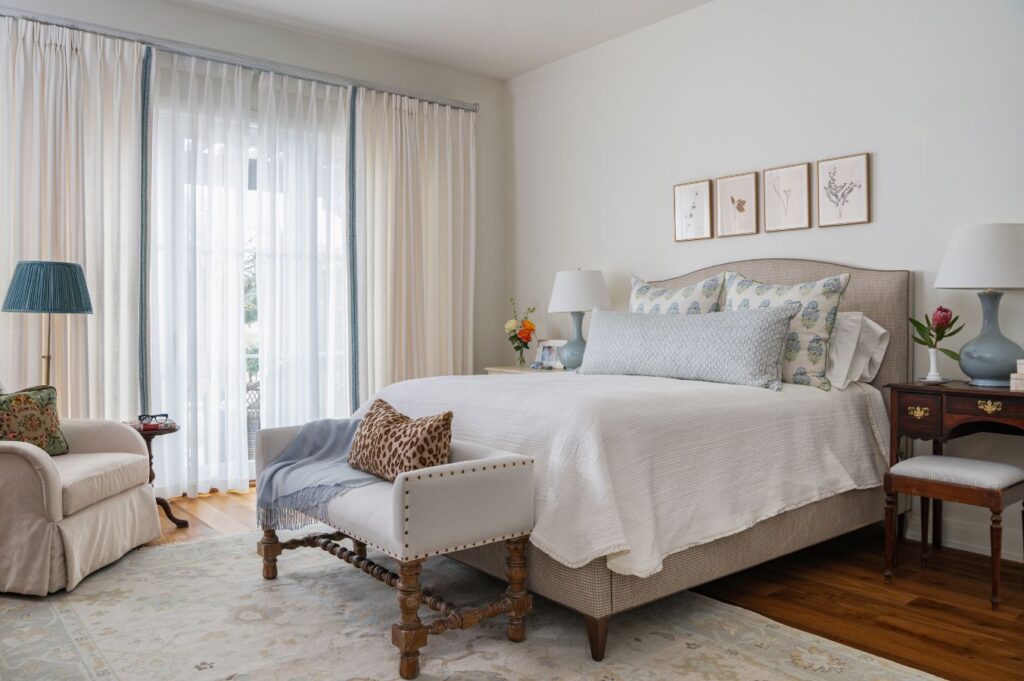 A bright bedroom with a large bed, white bedding, patterned pillows, a padded bench, an armchair, side tables with lamps, and framed art above the bed. Sheer curtains cover a window letting in natural light.