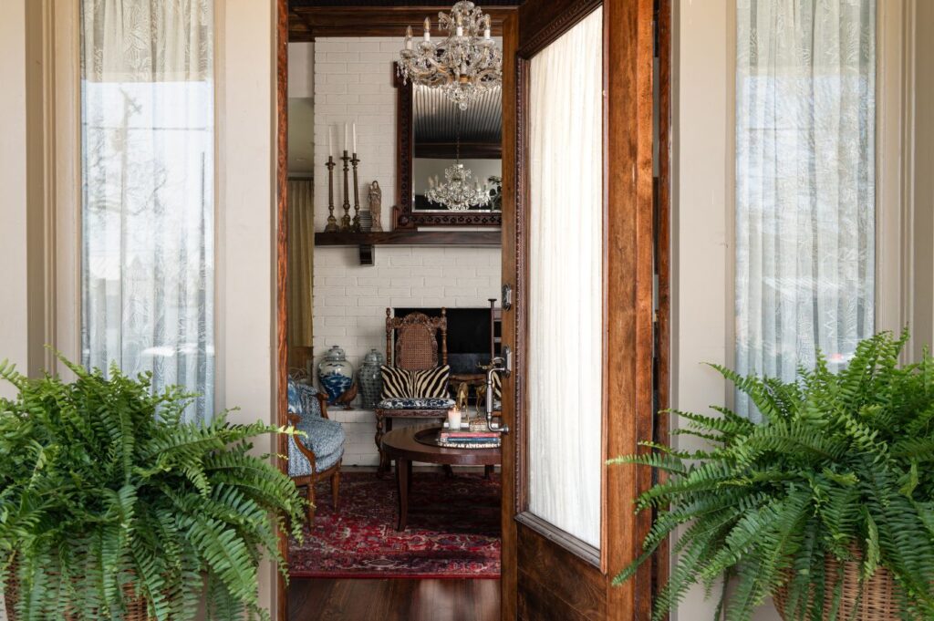 Open wooden door reveals a cozy living room styled by an interior designer Boerne TX, with a red ornate rug, antique chairs, round table, chandelier, wall mirror, white brick wall, ferns at the entry, and sheer curtains framing the doorway.