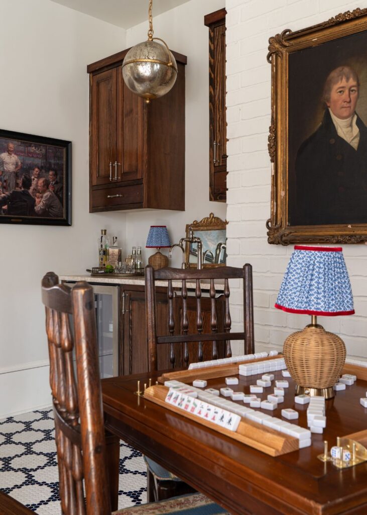 A cozy room by an interior decorator Boerne features a wooden table set for a tile game, rustic chairs, a small bar area, unique lighting, and paintings on the walls, including a portrait with a gold frame.