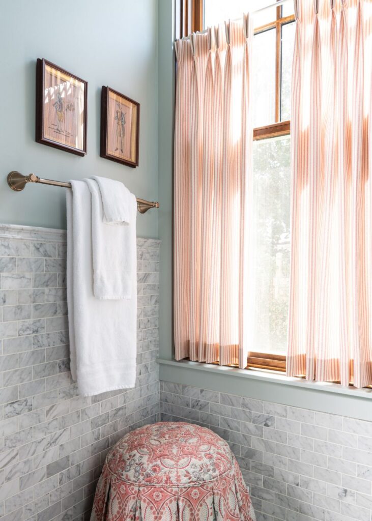 A bathroom corner by an interior designer in Boerne, TX, features a marble-tiled wall, towel rack with two white towels, framed pictures above, a window with striped curtains, and a round stool covered in red and white patterned fabric.