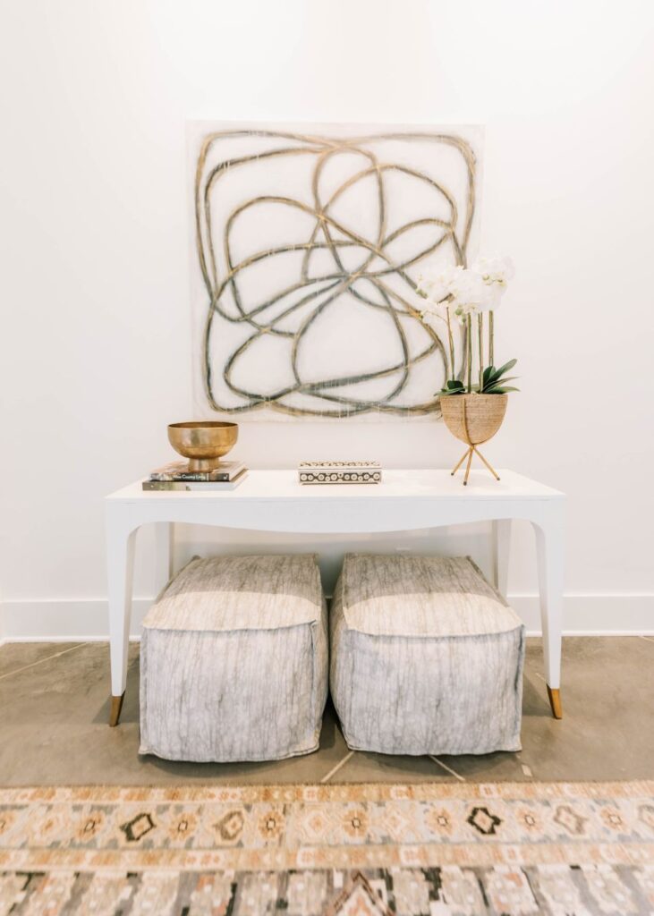 A modern white table with gold accents styled by an interior designer Boerne TX holds a potted orchid, decorative bowl, and books. Abstract wall art hangs above, with two gray textured ottomans placed on a patterned rug below.