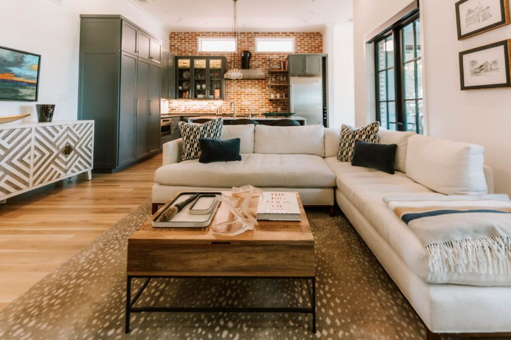 A modern living room with a white sectional sofa, patterned pillows, and geometric sideboard showcases the style of an expert interior designer Boerne TX. Light wood floors and large windows brighten the open kitchen with its brick backsplash.